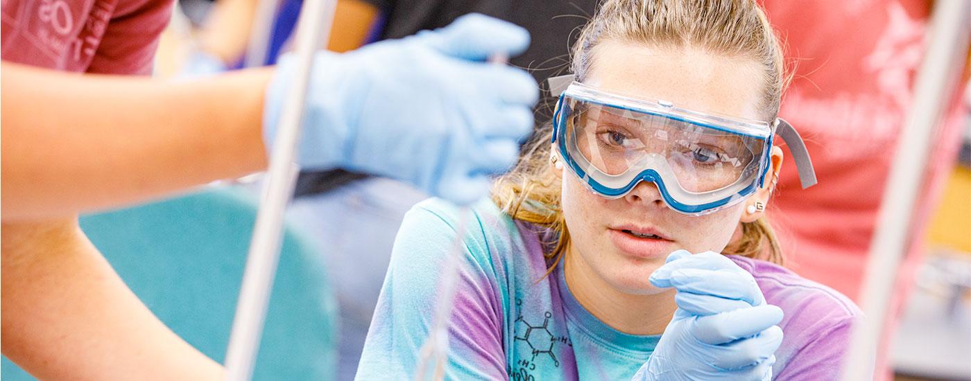 一名戴着护目镜的澳门十大赌城官方网站女学生在化学实验中观察另一名学生将液体放入烧杯中